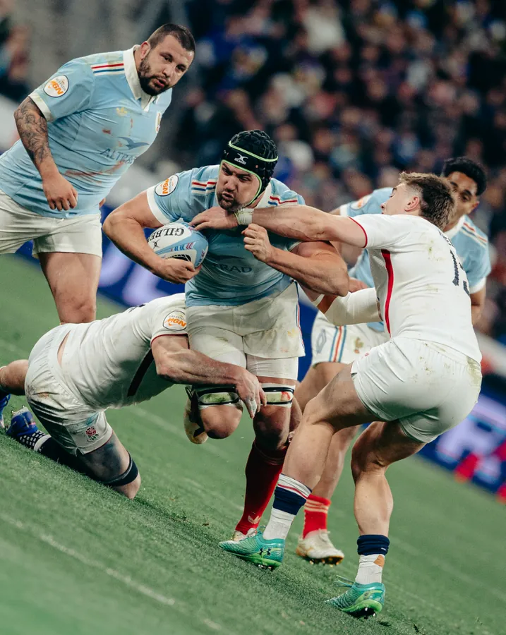 A rugby forward carrying the ball into two tacklers
